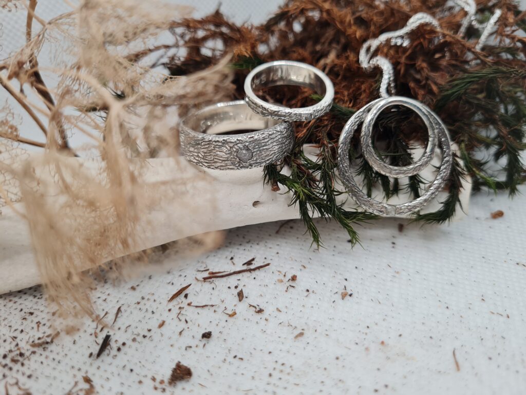 Twee zilveren ringen en een zilveren hanger rusten op takjes met groen. De sieraden hebben een ruwe afwerking.