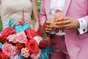 Een bruidspaar in een champagnekleurige jurk en een roze pak. De ene persoon houdt een kleurig boeket vast, de ander twee fruitige drankjes.