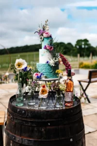 Een taart in drie lagen is versierd met bloemen en staat op een wijnvat omringd door bloemen in verschillende vaasjes.