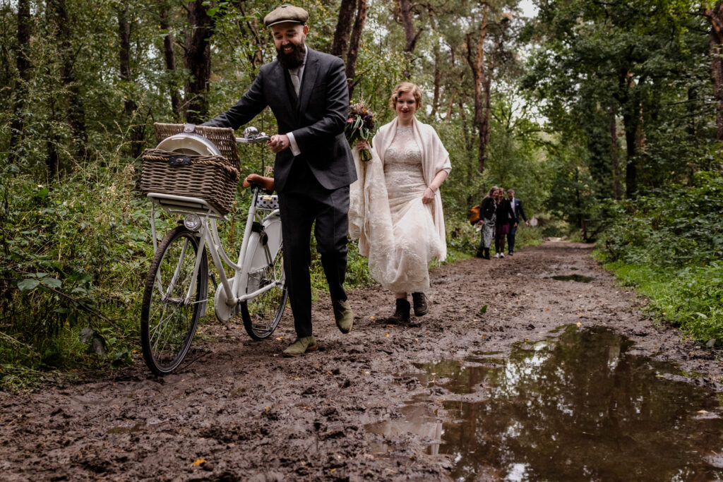 Een bruidspaar in vintage kleding loopt met een fiets over een modderig bospad