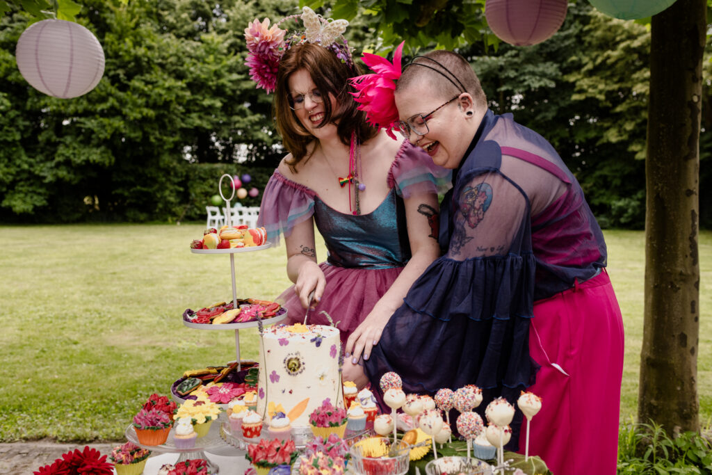Een non-binair koppel in kleurrijke outfits snijdt de taart op een fleurige sweet table aan.