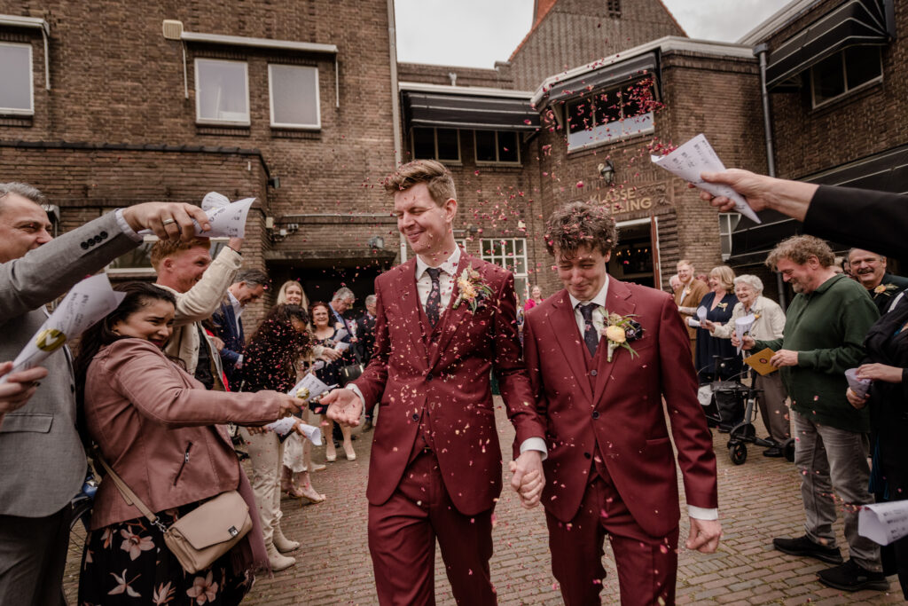 Twee bruidegommen in roodbruine pakken lopen tussen hun gasten door terwijl confetti gegooid wordt.