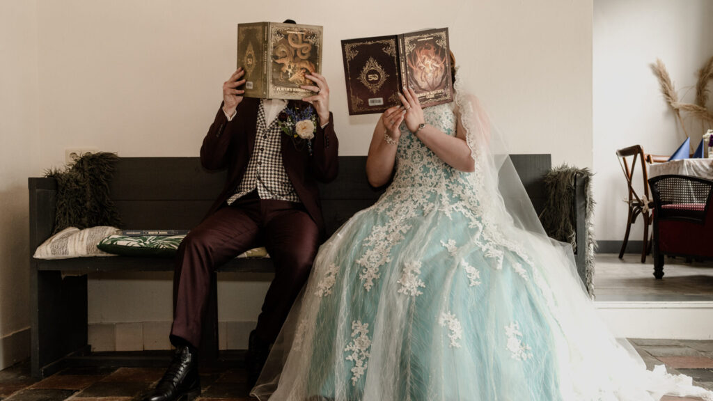 Bruidpaar in driedelig vintage pak en lichtblauwe jurk zit op een bankje met fantasy boeken voor hun gezicht.